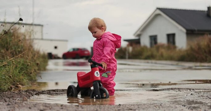 Small oneyear old baby girl  and mother playing in mud puddles after heavy rain. Jumping, throwing rocks and examine new . Wearing pink purple raincoat and Wellingtons. Riding a red bike, dogs around