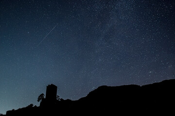 Nighttime photograph of a castle atop a mountain, with a starry sky and a shooting star in the sky....
