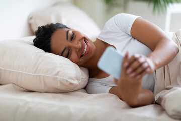 Obraz na płótnie Canvas Smiling Black Woman Using Smartphone While Lying In Bed At Home
