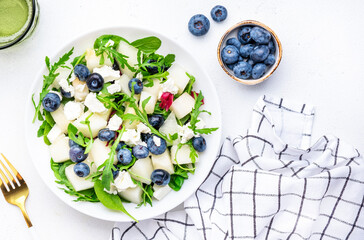 Delicious healthy salad with sweet melon, red onion, feta cheese, blueberries, arugula and mixed herbs, white table background, top view