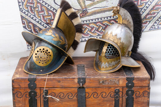 2 ancient roman gladiator helmets on a brown wooden chest with a mosaic image in the background