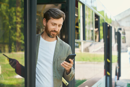 Stylish young man holding smartphone and getting off bus in city. Front view of busy bearded guy checking time on cell phone, while exiting of transport on bus stop. Transport, schedule concept.
