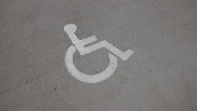 Handheld Slow Motion View Of A White Handicap Sign With Wheelchair Logo On A Concrete Cement Ground Near Parking Lot And Public Park