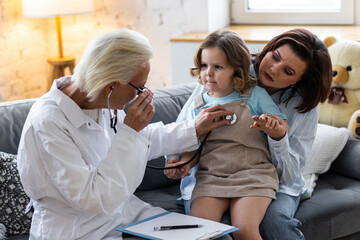 Obraz premium Concept healthcare medical assistance, insurance. Kind senior female paediatrician doctor examining little child sitting on mother's laps, during home visit or clinic check up. Prescribing treatment