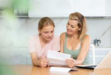 Fototapeta na wymiar Cheerful woman with teenage daughter getting good news. Happy family reading letter together while sitting at kitchen table with laptop at home..