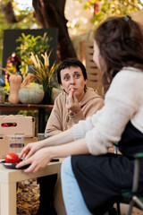 Cheerful woman tasting fresh homegrown veggies before buying produce and sitting at table with local farmers market vendor. People visiting organic food fair, enjoying products sample.