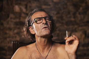 Fototapeta premium An older man, about 60 years old, of Serbian nationality with long hair and glasses, thoughtfully smokes a cigarette on a dark background. Artist, creative person. Happy face.