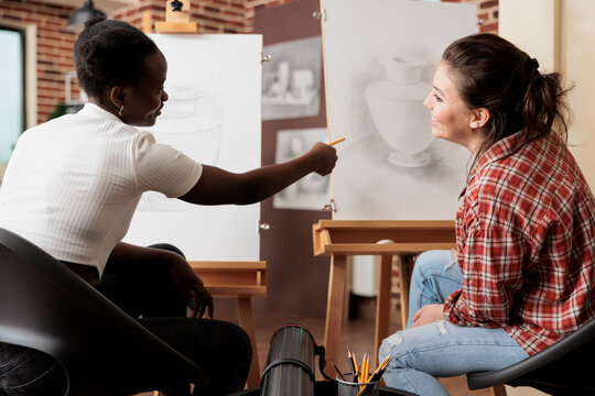 Two happy young women friends learning new things together, attending group art class, discussing artwork and smiling. Young diverse people talking communicating during drawing class for adults
