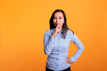 Portrait of filipino woman making silence gesture by putting forefinger over lips posing over yellow background. Silent woman shushing people indicating secrecy and confidentiality.