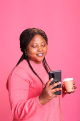 Smiling african american woman holding cup of coffee while browsing on social media, posting picture during lesiure time. Young adult watching funny video on smartphone whle posing in studio