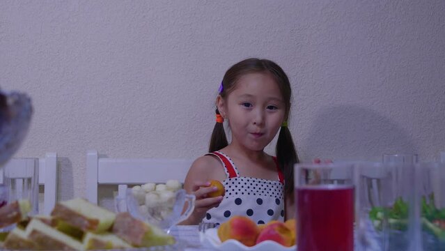 A cute girl picks up a fruit, points to the camera and eats