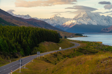 A car driving along a winding road beside a lake with snow capped mountains in the background.