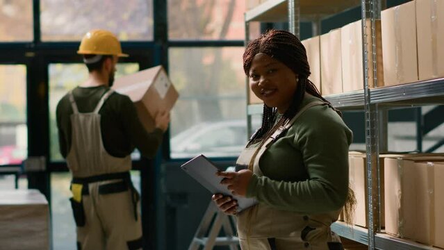 Portrait of joyful head of logistics in warehouse holding tablet, checking packaging while trainee in background handles packaged items in cardboard boxes ready to be shipped