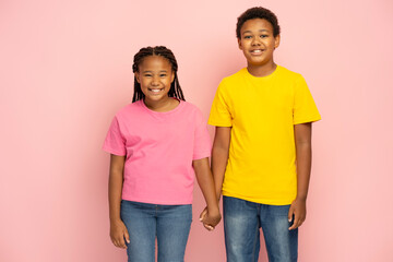 Fototapeta premium Young smiling African American kids, brother and sister wearing pink and yellow t shirts holding hands standing together isolated on pink background. Friendship concept