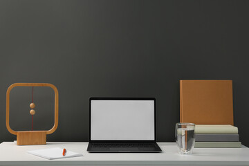 Stylish workplace with laptop, glass of water, books and decor on white table near grey wall