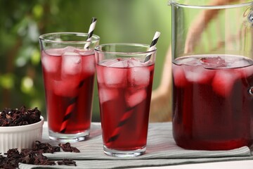 Refreshing hibiscus tea with ice cubes and dry roselle flowers on white table against blurred green background