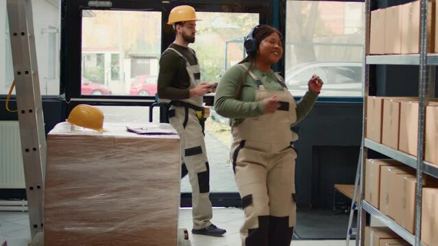 Happy warehouse sorter listening music enjoying herself while checking labels on cardboard boxes. Cheerful worker wearing heaphones and dancing around while walking through storage room rows