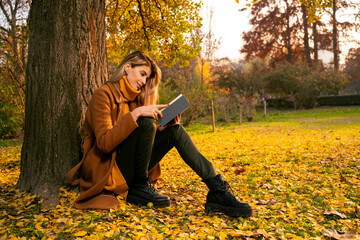 Pretty young woman sitting under a tree and reading a book, autumn season 