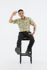 Naklejka premium Isolated cutout studio full body shot Asian vintage classic male fashion model with mustache neck arm tattoo wearing eyeglasses in casual outfit posing sitting on chair on white background
