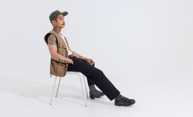 Portrait isolated cutout studio full body shot Asian vintage classy mustache neck arms tattoos male fashion model in casual fashionable safari vest and cap sitting on white chair on white background.