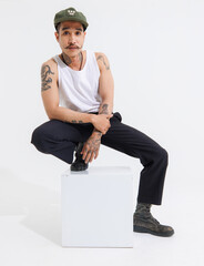 Isolated cutout studio shot Asian vintage classy mustache with neck arms hands tattoos male fashion model in casual fashionable sleeveless shirt cap boots sitting on square box on white background