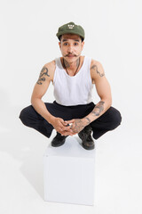 Isolated cutout studio shot Asian vintage classy mustache with neck arms hands tattoos male fashion model in casual fashionable sleeveless shirt cap boots sitting on square box on white background