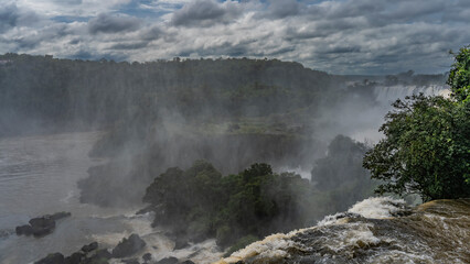 A stream of seething water collapses from a ledge. Spray and mist in the air. Lush tropical vegetation all around. Clouds in the sky. Argentina. Iguazu Falls.