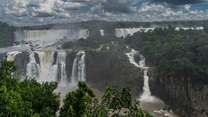 Fototapeta premium Waterfall landscape. Streams of water collapse from the ledges of rocks and flow along the rocky riverbed. Lush tropical vegetation all around. Clouds in the sky. Iguazu Falls. Brazil.