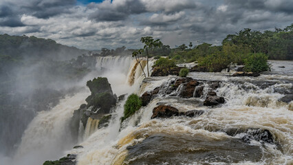 A stream of swirling water rushes along the rocky riverbed and collapses into the abyss. Green vegetation on the stones. Spray, fog all around. Clouds in the sky. Iguazu Falls. Argentina.