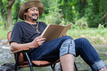 Obraz premium Happy bearded Asian man sitting alone in a chair near a stream in the countryside and enjoying the forest scenery while reading a book.