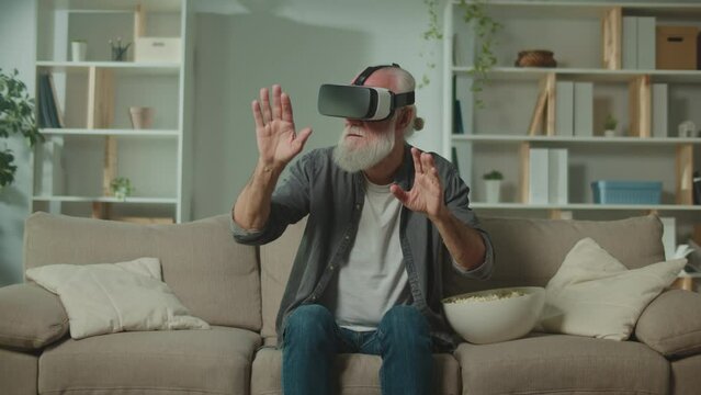 A Modern Old Man in VR Glasses Examines and Touches Everything Around. An Elderly Man in VR Glasses Sitting in the Sofa with a Popcorn.Virtual Reality Education for the Elderly.