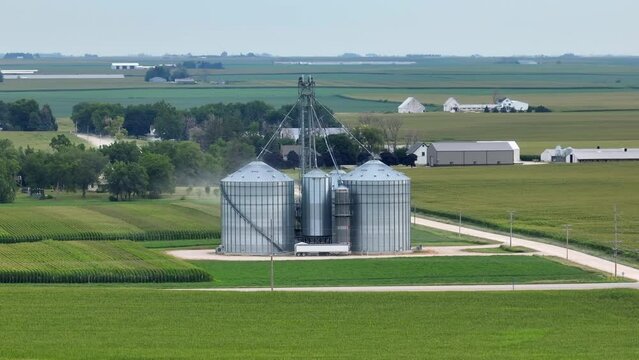 Wide orbit shot of grain bins in flat, sprawling midwest USA. Corn and grain fields for miles. Long aerial zoom shot of grain elevator in summer.