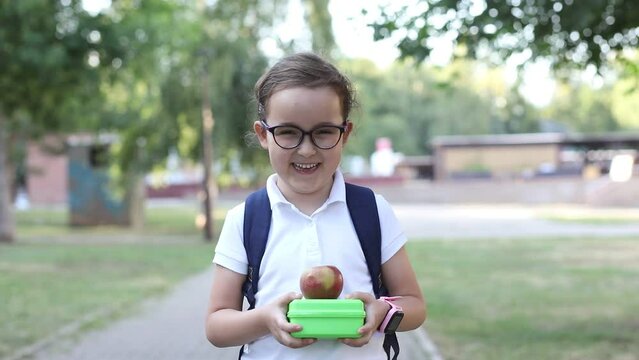 Cute little schoolgirl with backpack holding in a hands lunch box. Near by school outdoor. Food for kids