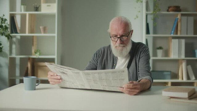 A Serious, Smart Old Man Sits at a Table and Reads a Newspaper.An Elderly Man with a Newspaper Alone at Home, Sees the News and Current Events in the Newspaper.Daily Newspaper Concept