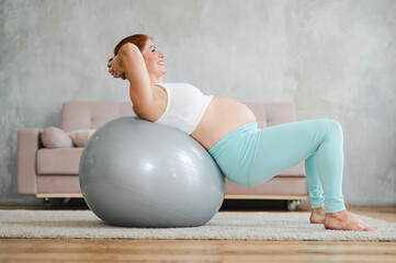 Pregnant red-haired woman doing fitball exercises at home. 