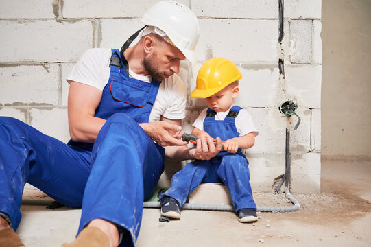 Male builder showing wire stripper cutter tool to child. Man and kid in safety helmets and work overalls sitting by brick wall in building under construction. Home renovation and parenting concept.