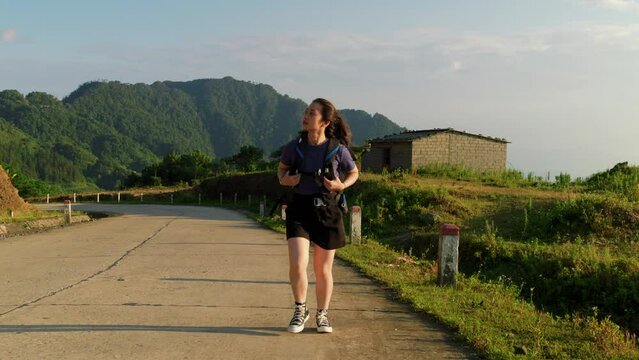 Frontal tracking view of asian woman carrying hiking backpack across countryside pathway at sunset