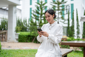 A cute and happy Asian woman using her smartphone while sitting at a bench in a garden.