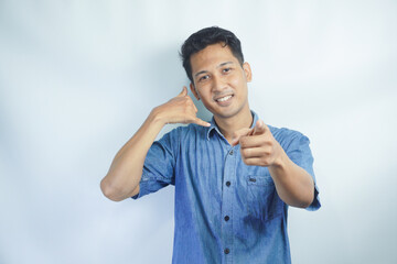 Fototapeta premium Young Asian man wearing blue shirt standing over isolated white background smiling doing talking on the telephone gesture and pointing to you. Call me.