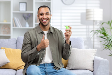 Portrait of a young African American man sitting on the couch at home and holding a throat spray in his hands and showing a super gesture with his finger - obrazy, fototapety, plakaty