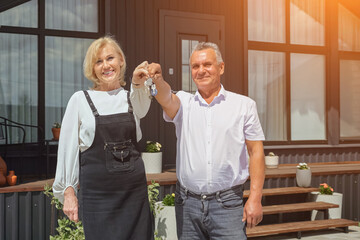 Mature man and woman smile holding keys to new vacation home. Family become new owners of cottage residence from estate agency, sunlight