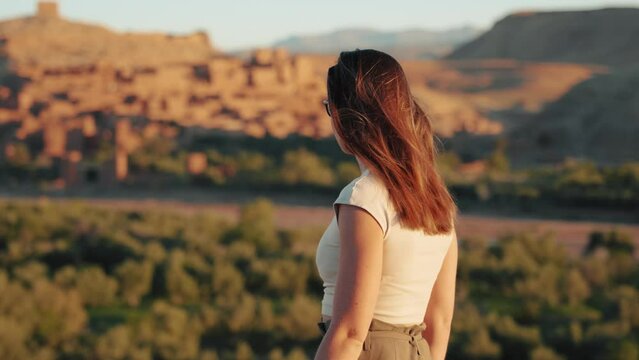Close view of young caucasian woman walking and looking around through desert landscape at sunrise in Ait Ben Haddou, Morocco.
