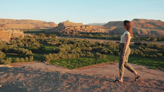 Wide view of young caucasian woman walking and looking around through desert landscape at sunrise in Ait Ben Haddou, Morocco.