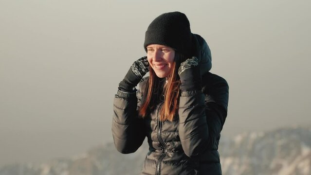 Young caucasian woman in beanie adjusting her winter jacket during sunrise on top of a mountain.