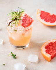 A soft pink glass with an iced grapefruit drink, garnished with sugar and a sprig of rosemary, around a piece of grapefruit.