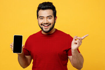 Impressed happy young Indian man wear red t-shirt casual clothes hold in hand use mobile cell phone with blank screen workspace point aside on area isolated on plain yellow orange background studio - obrazy, fototapety, plakaty