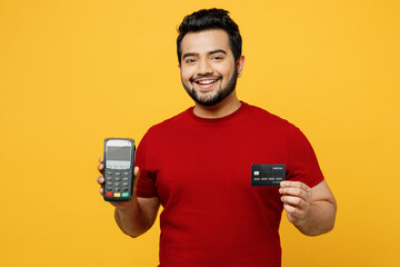 Smiling young Indian man he wearing red t-shirt casual clothes hold wireless modern bank payment terminal to process acquire credit card isolated on plain yellow orange background. Lifestyle concept.