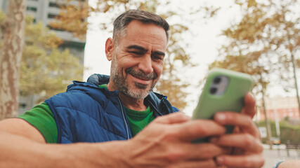 Fototapeta premium Close-up,happy mature man wearing casual clothes sitting on bench looking through information on smartphone