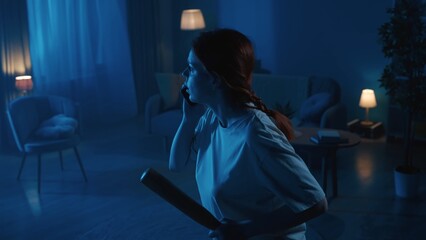 Shot of a young woman going around a dark room in search for burglars, intruders, thieves. She holds a baseball bat and a phone to call the emergency hotline.
