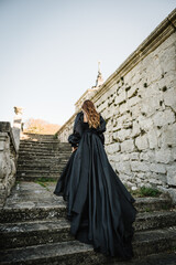 Fototapeta premium Female in long black dress walk on stairs near ancient palace at sunset. Model girl goes in nature autumn day. Luxury woman near old Pidhirtsi Castle, Lviv region, Ukraine. Stylish bride. Back view